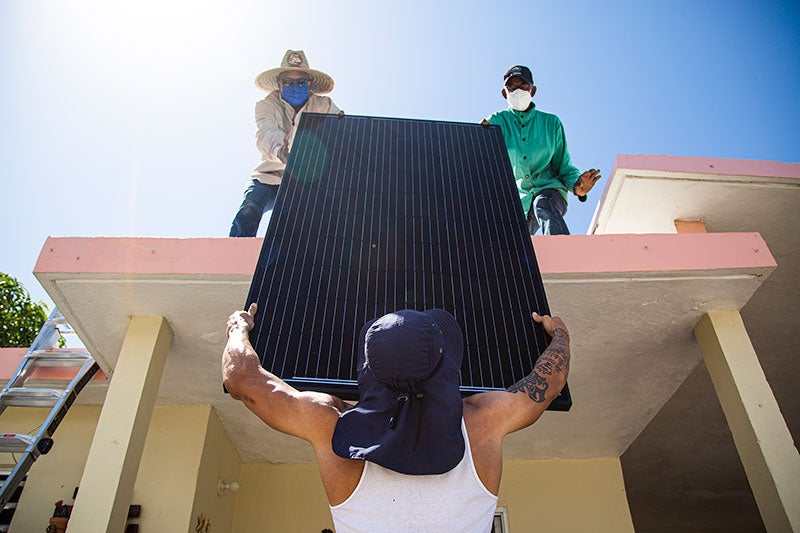 Solar panel lowered from roof by two volunteers to volunteer below. Person below has hands on both sides of the panel. People on roof are guiding the transfer. All are wearing hats. The sun is bright. Face of the two on the roof are visible - they are wearing masks.