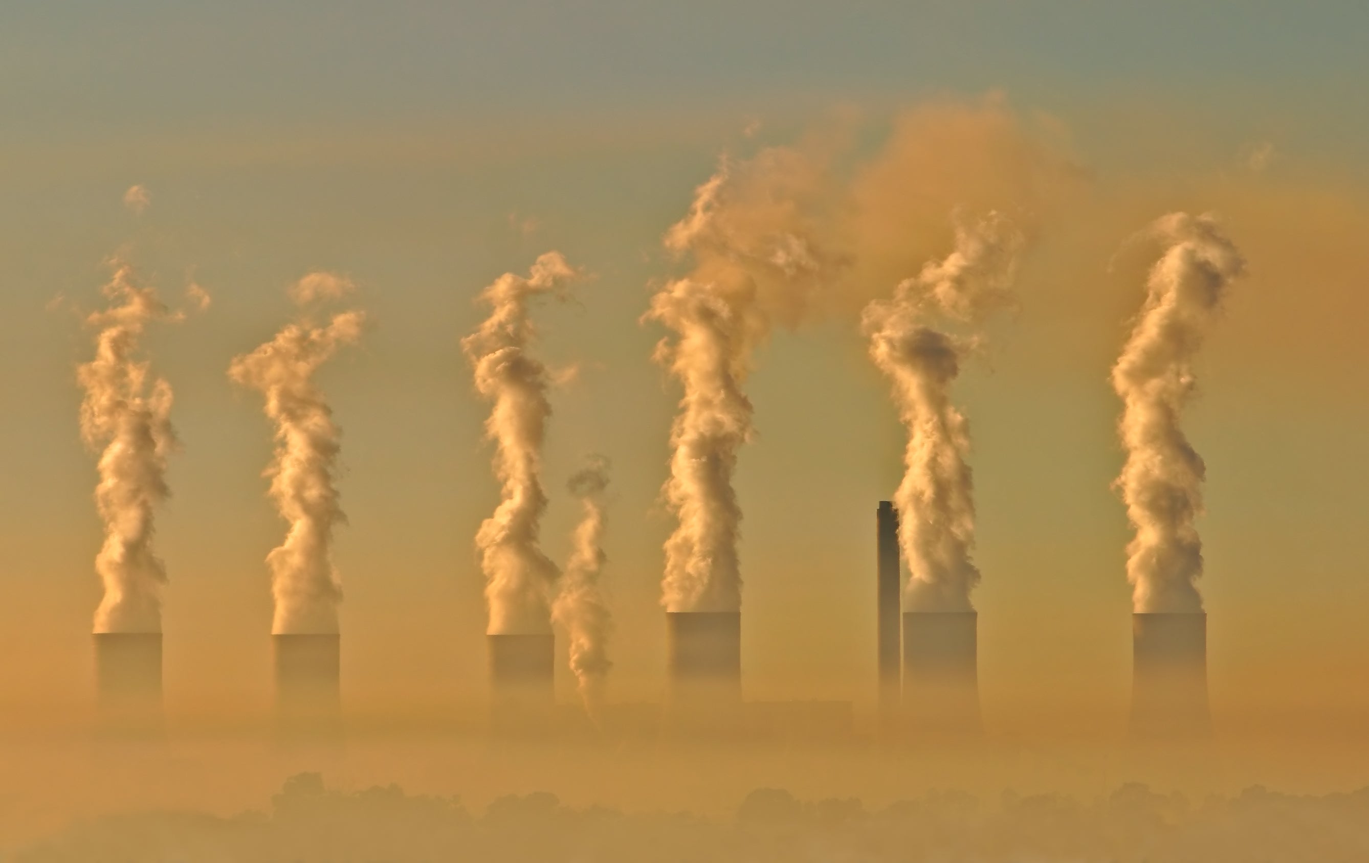 Smokestacks loom up from a hazy sky, tinged with orange, at sunset. Smokestacks loom up from a hazy sky, tinged with orange, at sunset.