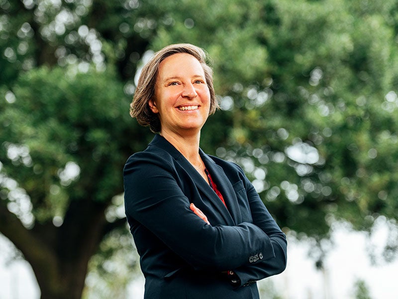 Jen Powis, Managing Attorney of the Earthjustice Gulf Regional Office. Jen Powis, Managing Attorney of the Earthjustice Gulf Regional Office stands smiling in blue jacket with arms crossed in front of a verdant tree.