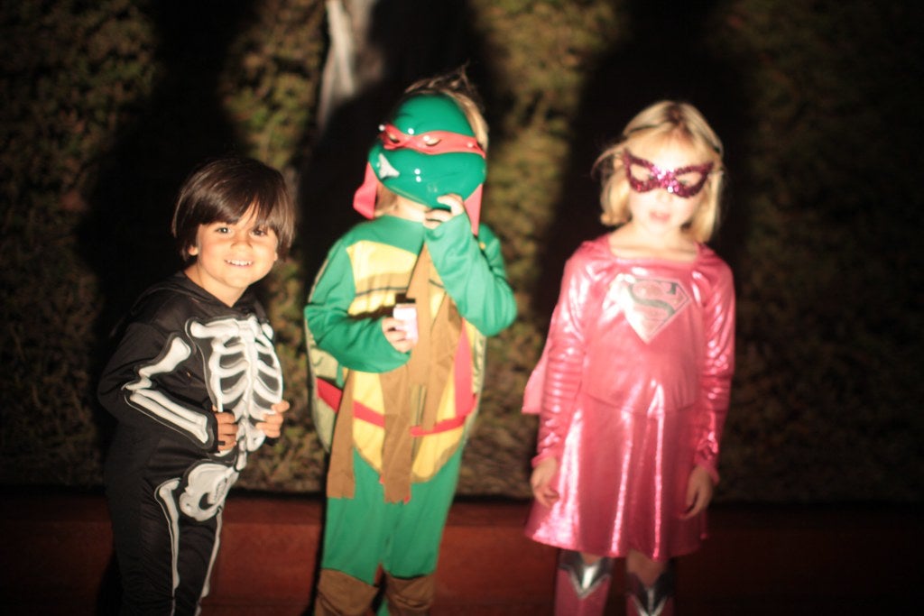 Three children dressed for Halloween in, from left to right, skeleton, Teenage Mutant Ninja Turtles, and Super Girl costumes.
