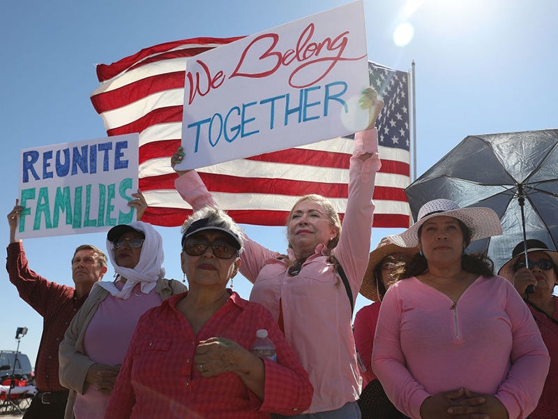 Activists stand in front of a U.S. flag, holding signs that say "We Belong Together" and "Reunite Families" in protest of family detentions outside of the Tornillo Port of Entry in Tornillo, TX.