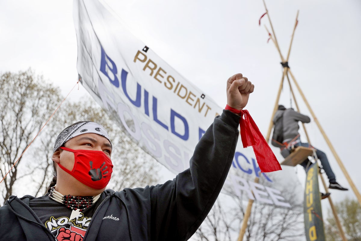 Photo of an Indigenous activist with fist raised in the air before a sign that reads "President Biden, build back fossil free."