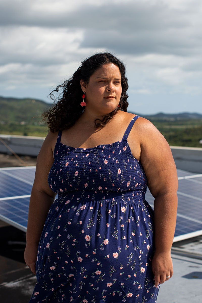 Amy Orta-Rivera standing. Solar panels behind her. Green hillscape behind solar panels. Clouds in the sky. She is wearing dark blue summer dress with thin straps. Sun shining on her.