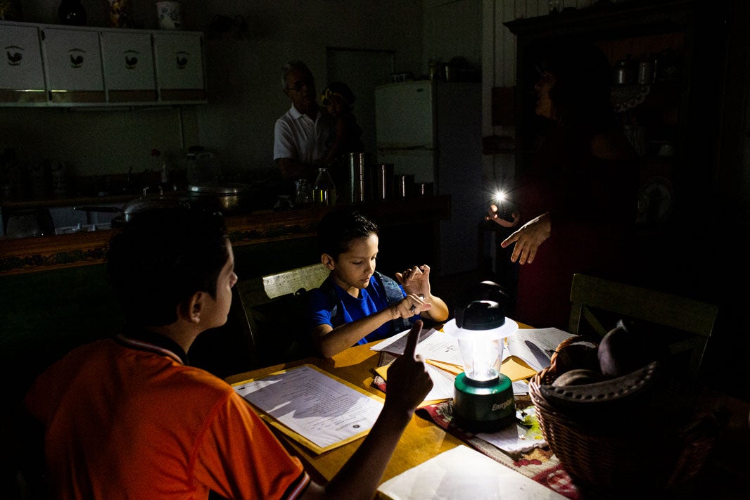 People gathered in a dim kitchen. Two youth sit at dining table. Paperwork and portable lamps sit beside basket of produce. Barely visible adults in background -- one holding a cellphone, shining the cellphone light onto a document in front of one of the youth.