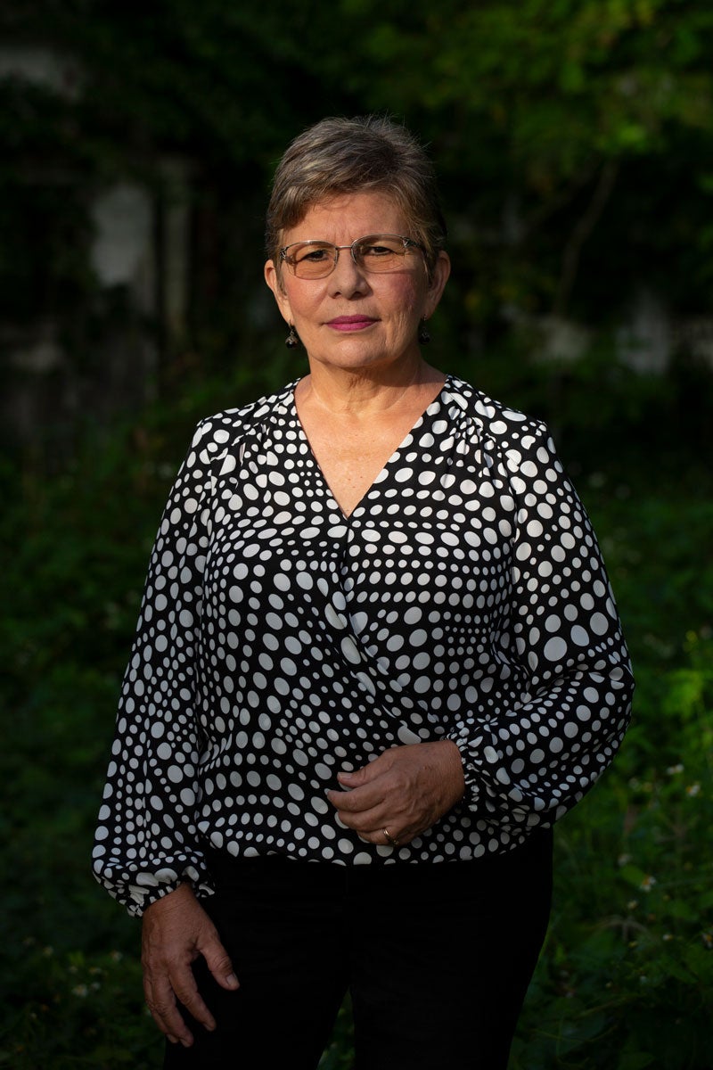 Myrna Conta stands outside. Blurred background is mostly green with foliage from trees and from the ground. She is wearing glasses, a black blouse with white dotted patterns, and black pants.