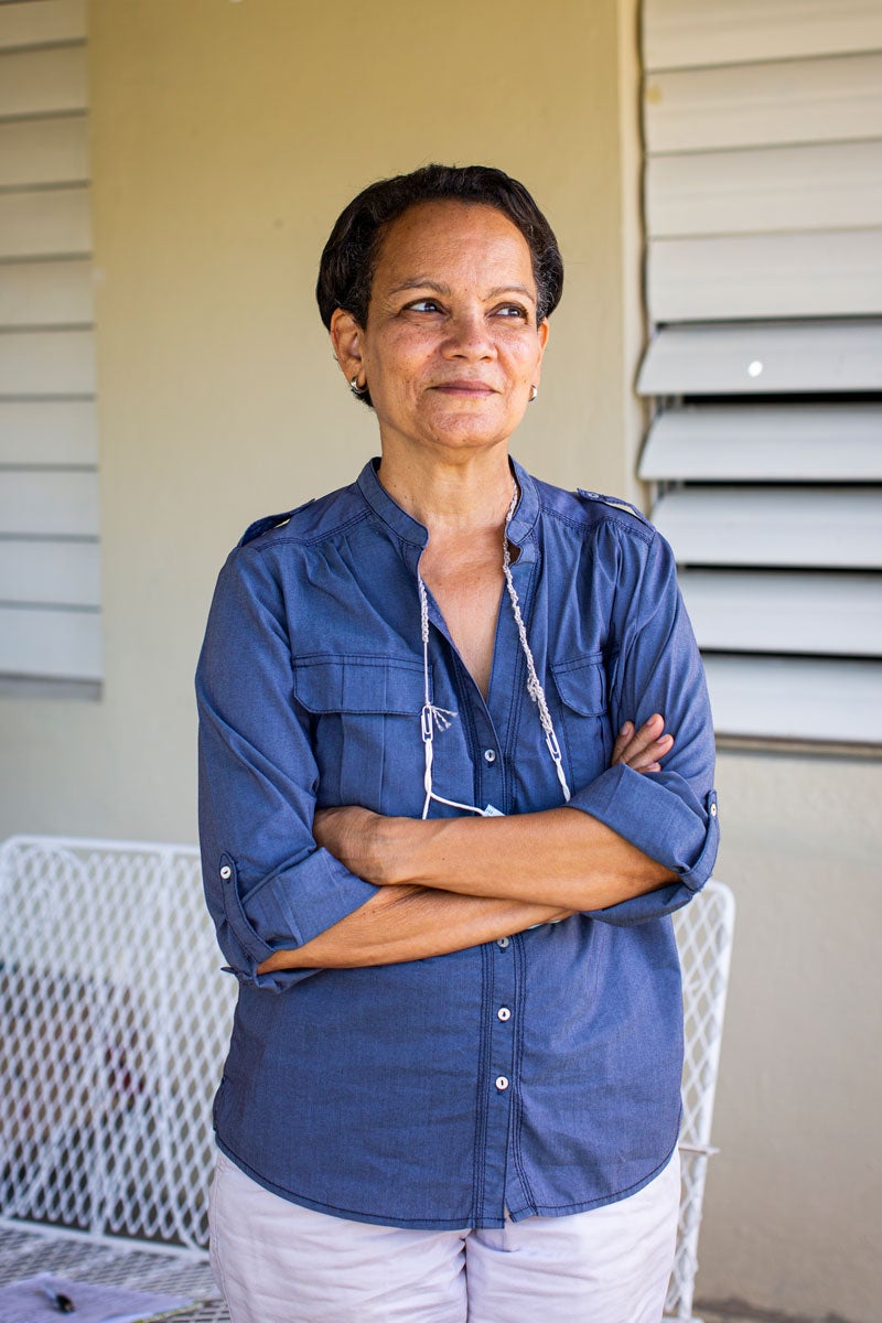 Ruth Santiago stands with arms crossed, smiling. Behind her is a beige wall with white blinds. In front of the walls is a white metal bench. She is wearing a blue denim blouse and a lanyard around her neck.