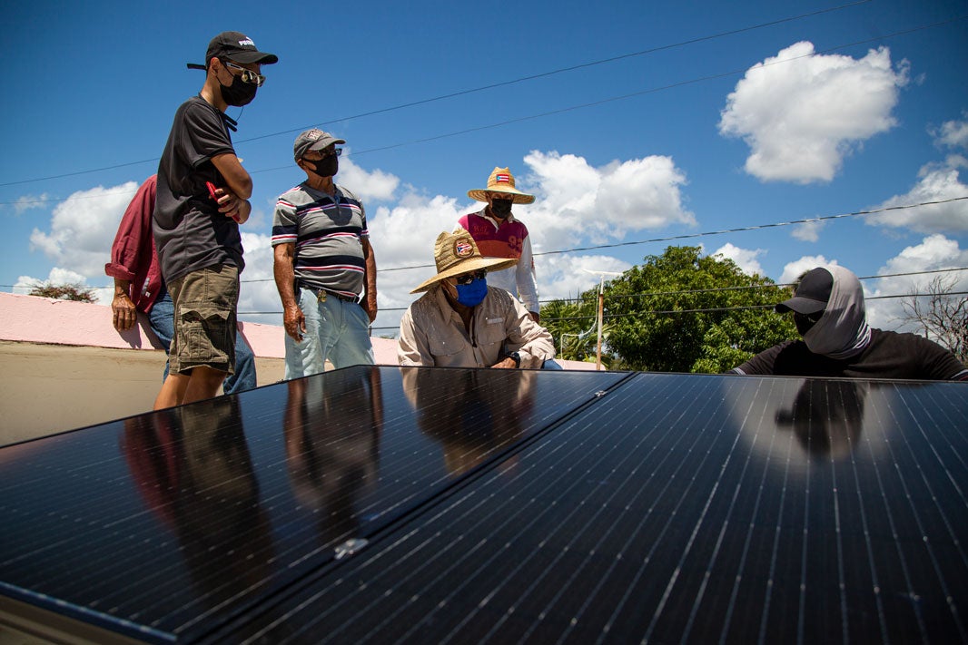 A group of six stand around a set of solar panels. They have sunhats on, sunglasses, and masks. One person with a fabric wrapped around his neck, face, and back of his head is crouched near the back of the panels. Blue skies, wispy clouds, sunny.