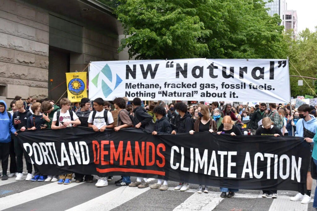 Young people on the street holding signs that say "Portland Demands Climate Action" and NW "Natural" Nothing Natural about it.