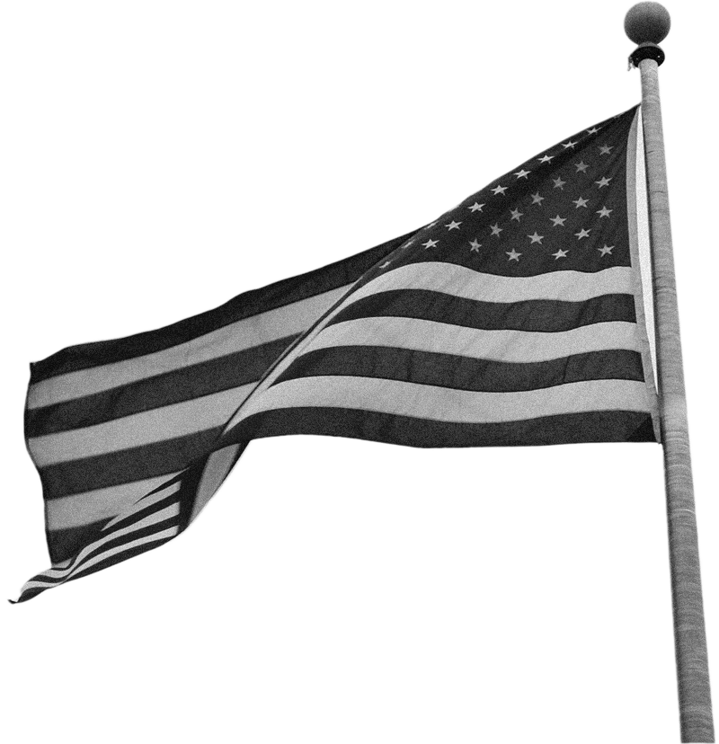 Black and white image of the United States flag, flying at the top of a flag pole in a slight breeze.