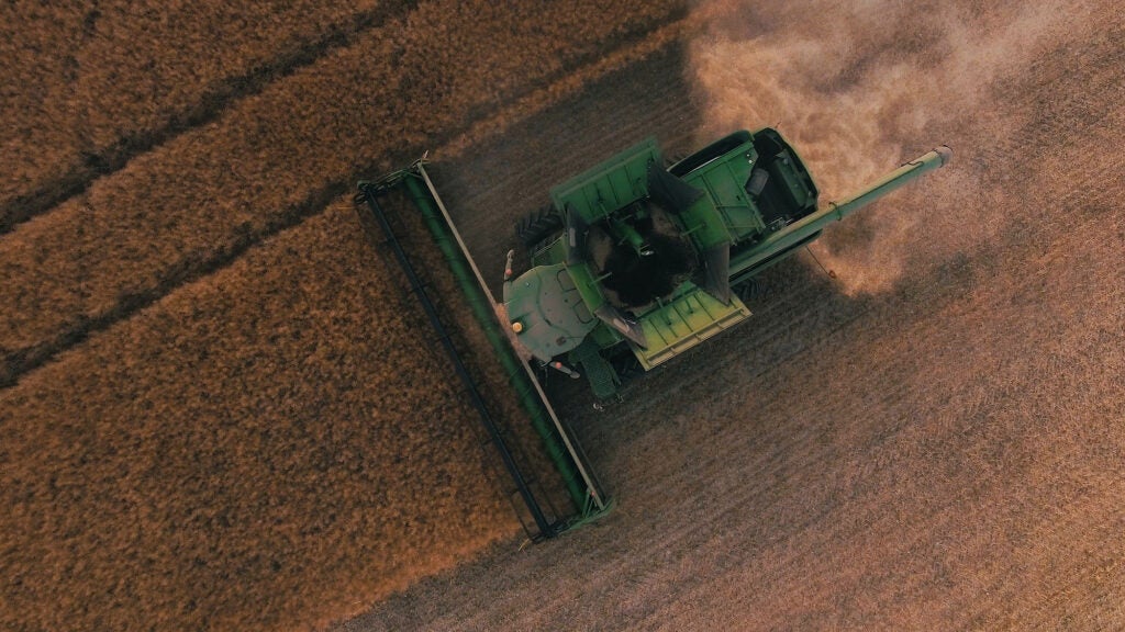 Aerial view of green mechanical combine harvesting in a brown field, with a dust cloud trailing behind the machine.