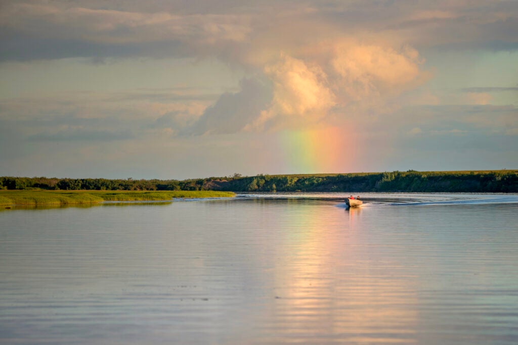 a small boat with several people on a wide, smooth river with a rainbow in the background.