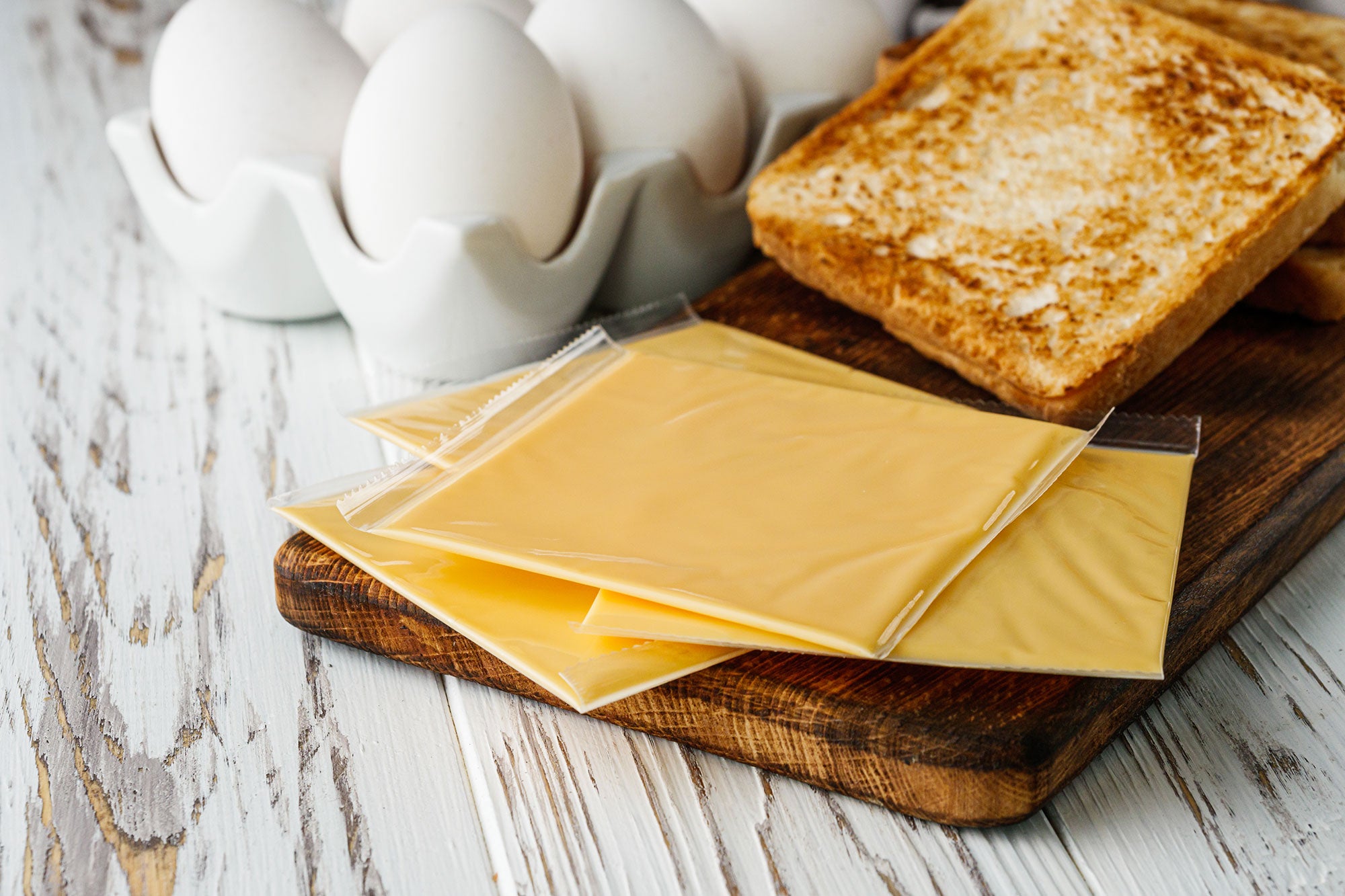 Sliced American cheese in plastic packaging are spread out on a wooden cutting board, next to toasted bread and white eggs in a ceramic egg-holder.