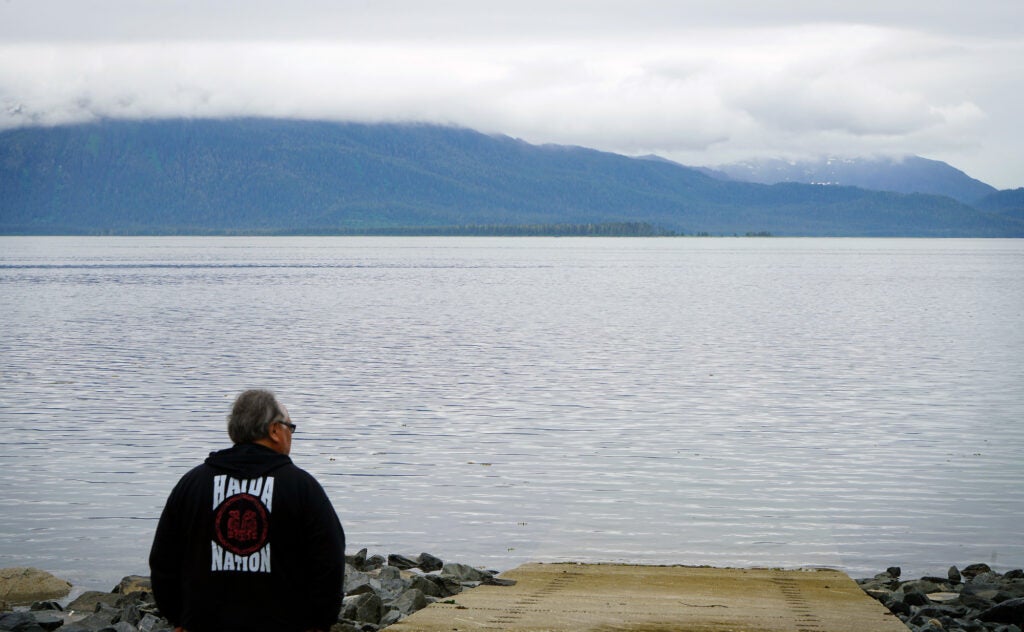 A man looks out at calm water on a cloudy day with mountains in the distance.