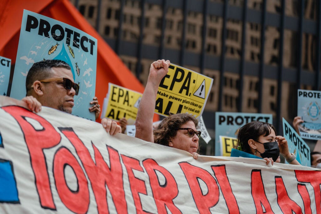 A line of people hold a long banner with the large words, "Make Power Plants Clean Up All Their Coal Ash" in front of a skyscraper in Chicago. Many more people are holding signs behind the banner with the words " Protect Lake Michigan" and EPA: Make Power Plants Clean Up All Their Coal Ash." A woman holding up the center of the banner stands with a raised fist.