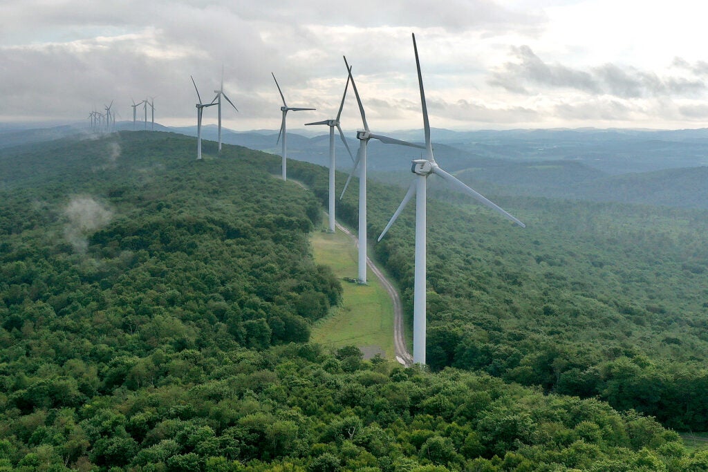 A long line of wind turbines on a tree covered mountain ridge.