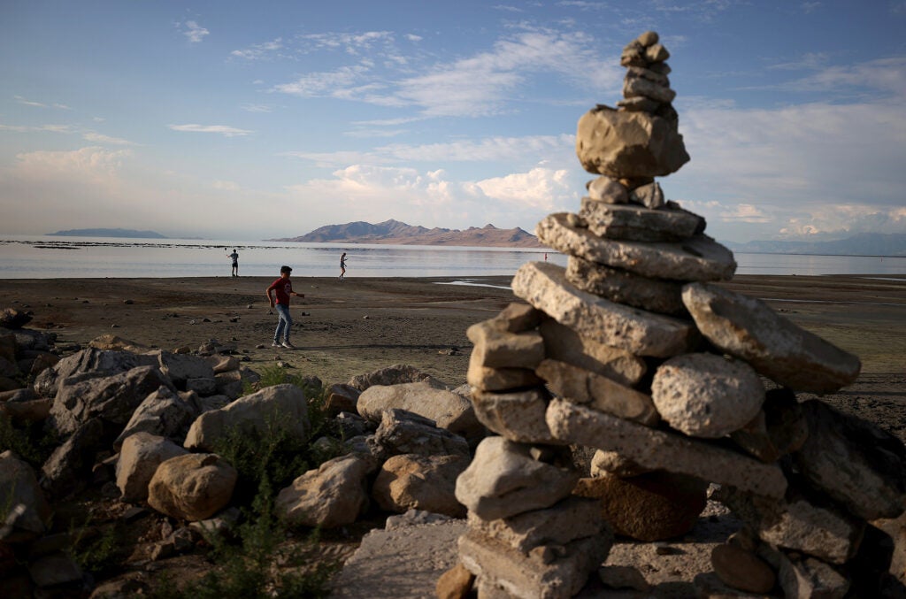 A stack of rocks on a large beach with people playing near the lake in the far distance.
