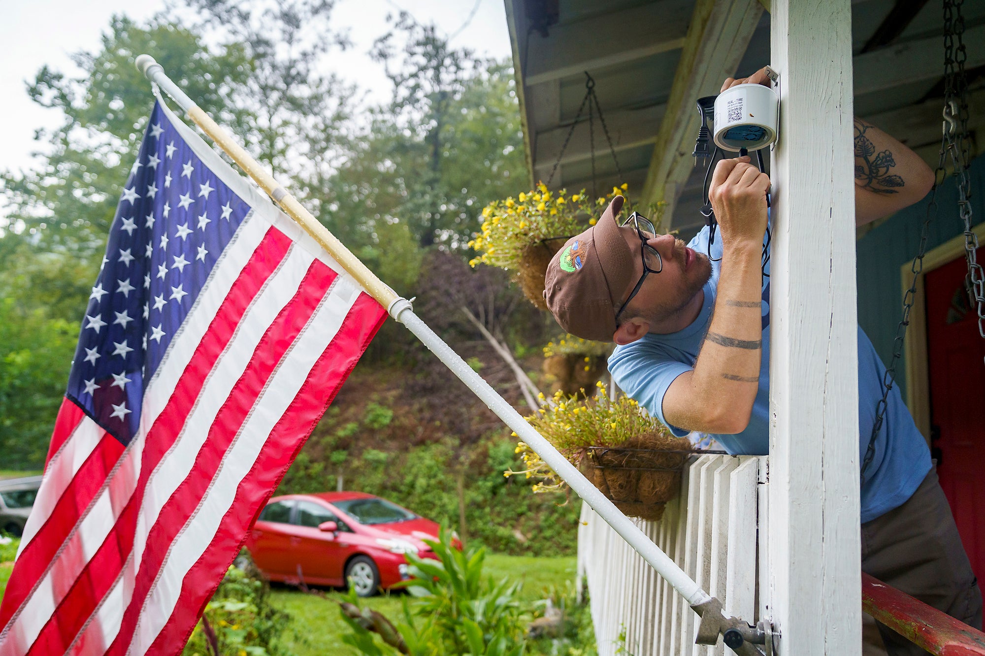A man putting a small white monitor on the front porch of a home with a large American flag on it.