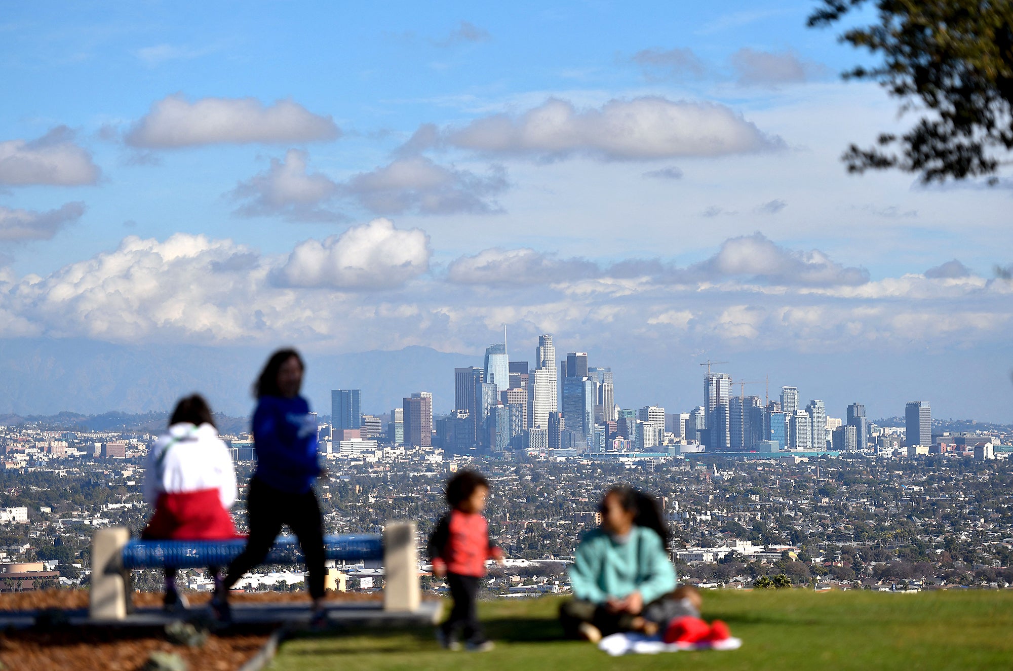 Out of focus people in the foreground with the Los Angeles skyline behind them, in focus, on a sunny day with clouds in the sky.