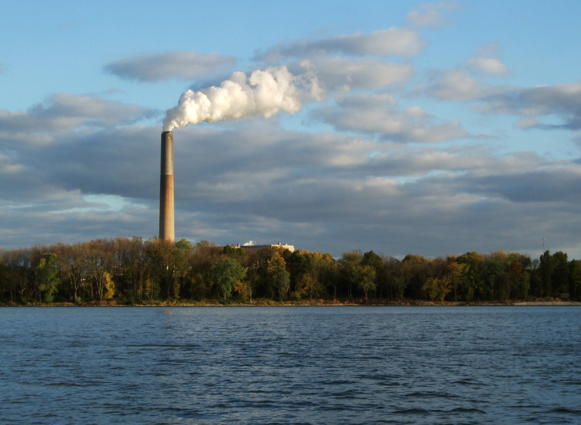 A tall smoke stack with plumes of smoke coming out of it next to a calm lake surrounded by trees.