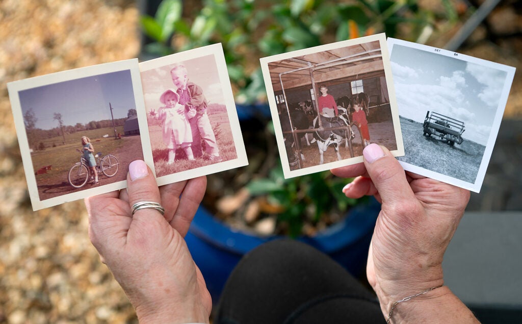 Four old color and black and white photos of farm life, including a girl on a bicycle, a toddler and young boy, kids with cows and a tractor, being held by Lori Phillips.