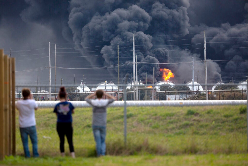 people in the foreground look at a chemical facility in flames with a sky full of dark smoke rising up.