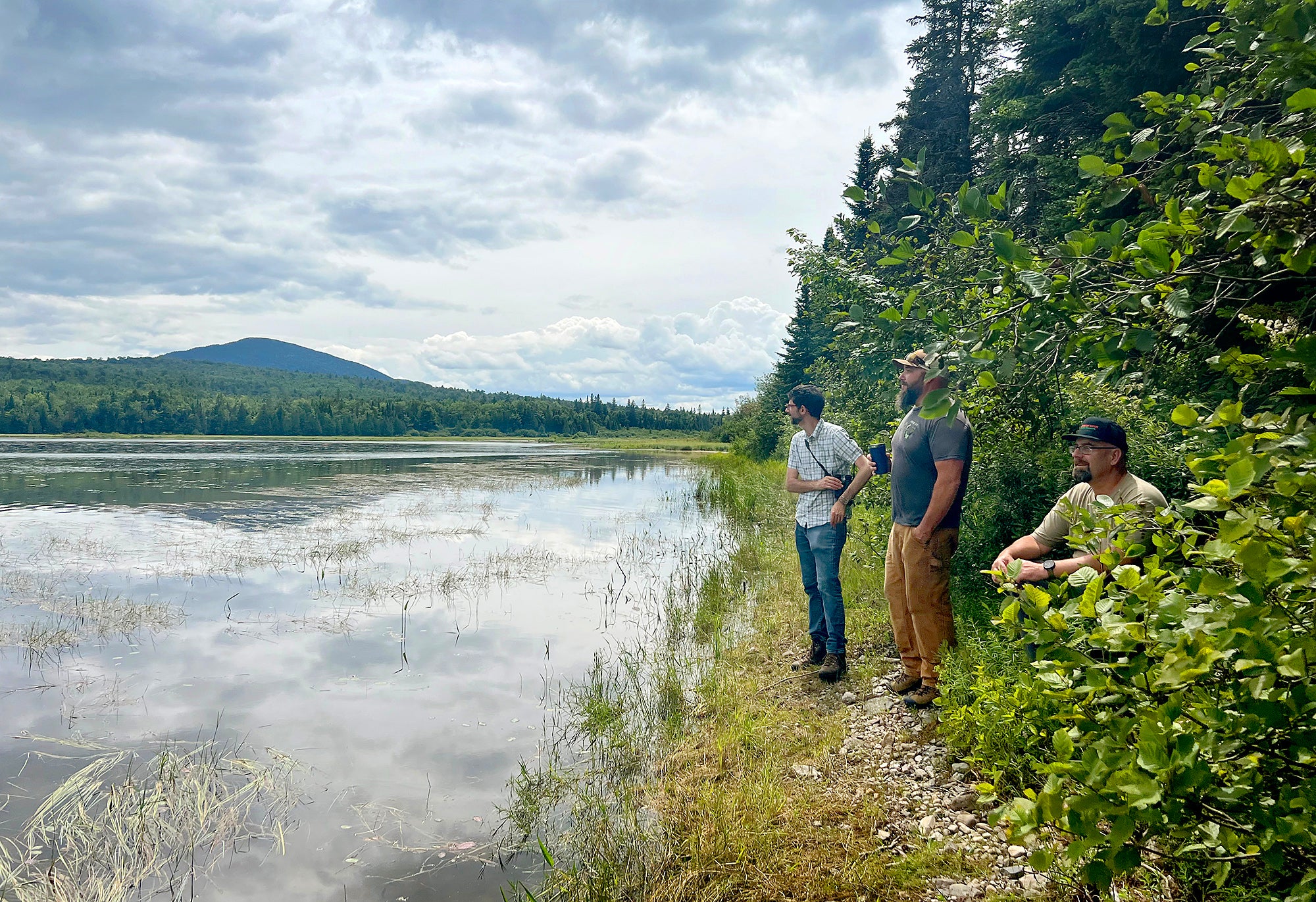 Three men stand next to a pond or river in a wooded area with a mountain in the background.