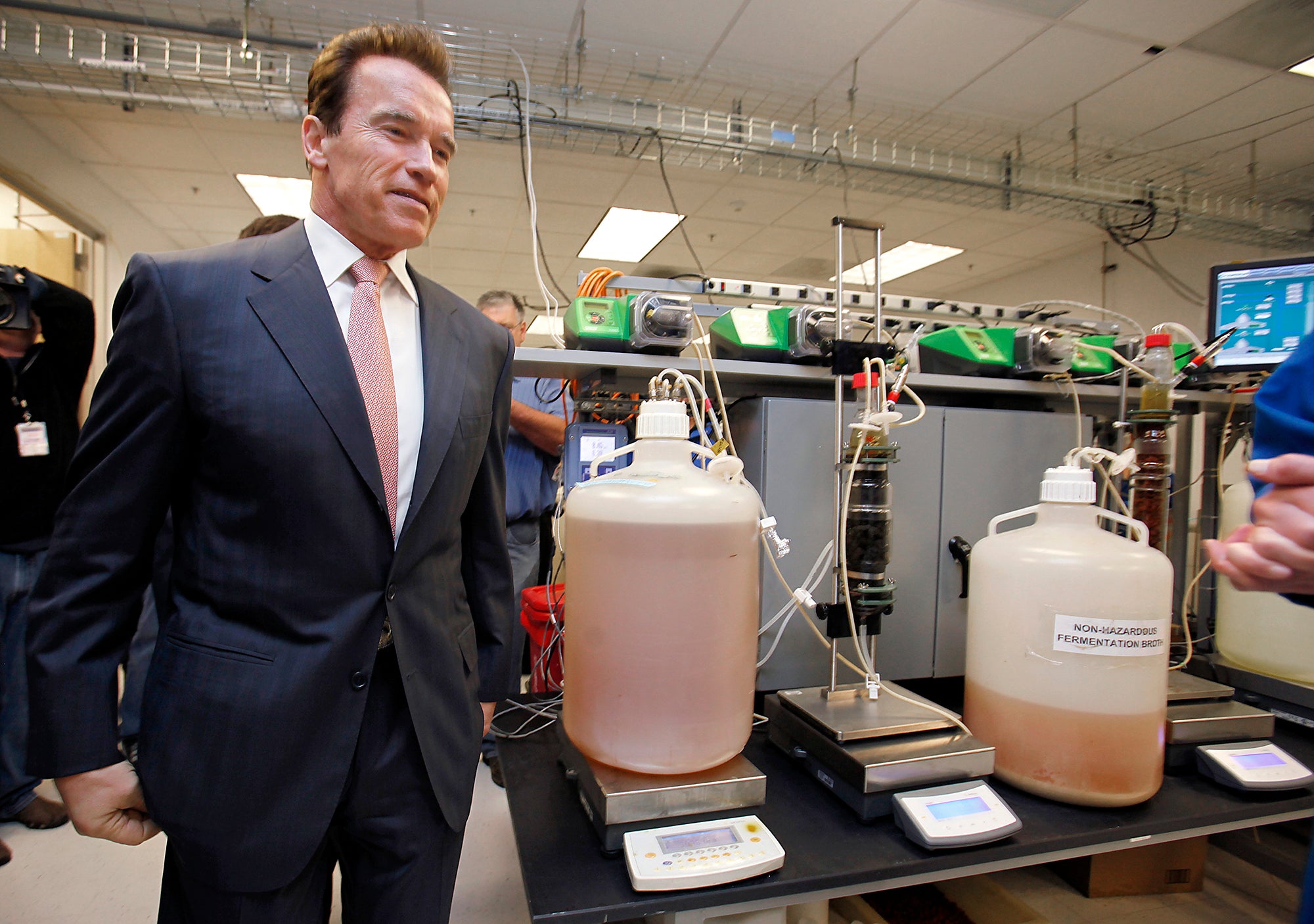 Arnold Schwarzenegger smiles and looks at two large jugs of brown liquid in a laboratory.