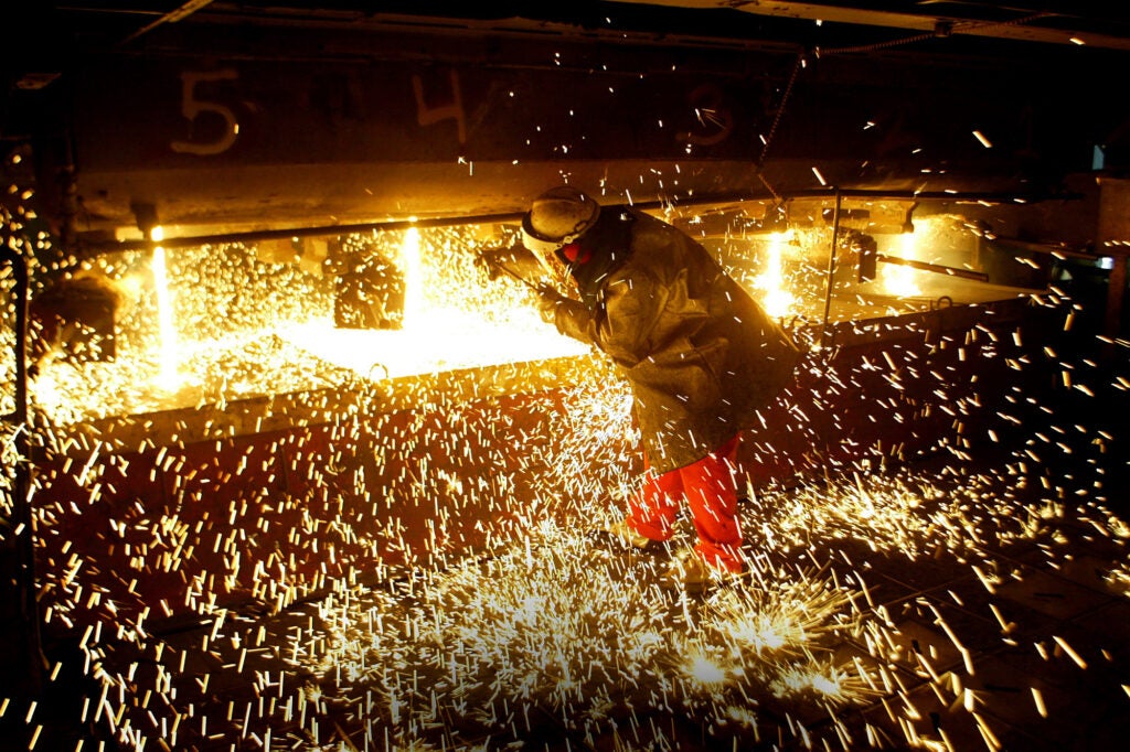 A person uses a rod in a machine filled with yellow melted steel with sparks flying around.