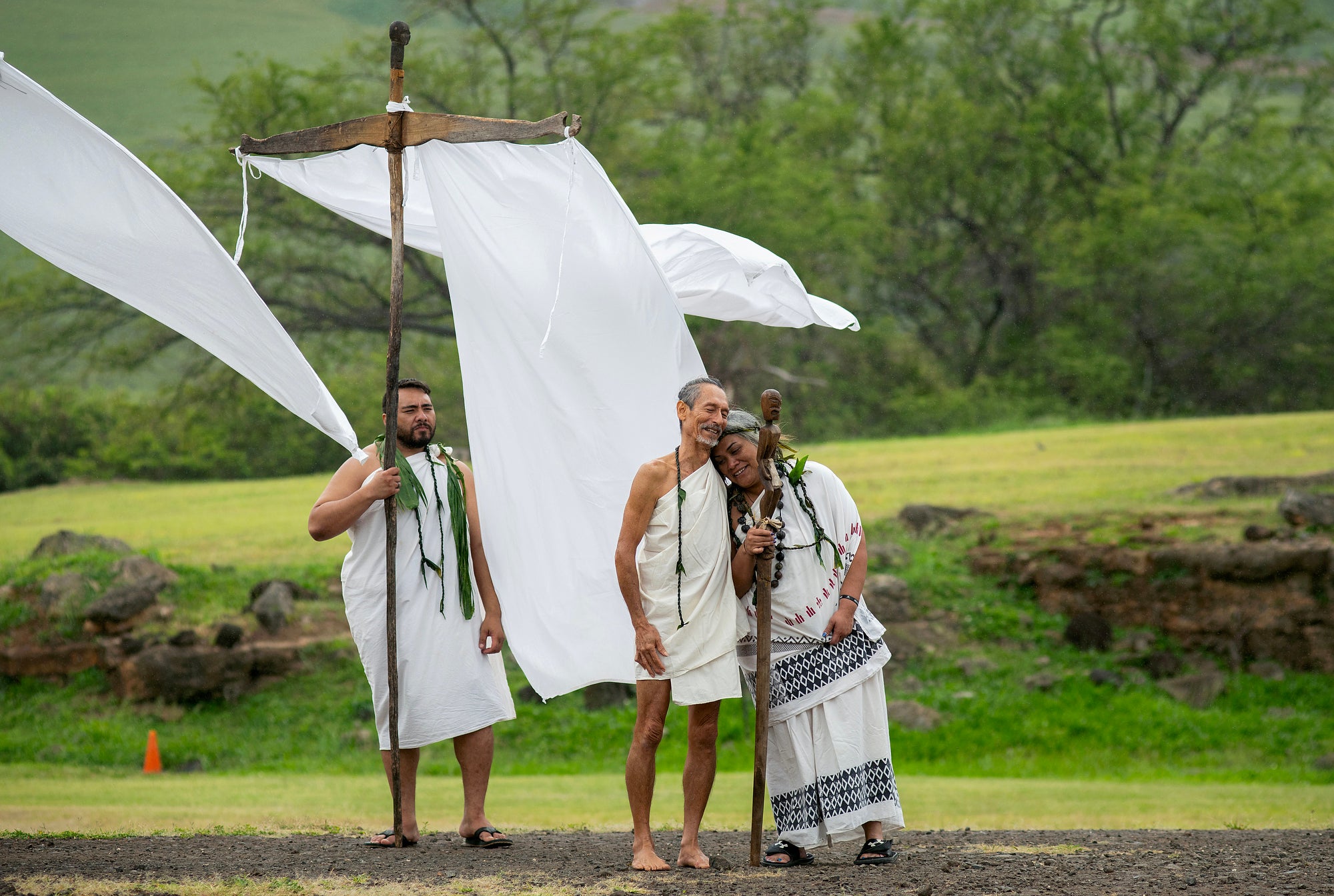 Three people in white traditional clothes stand in a green valley.