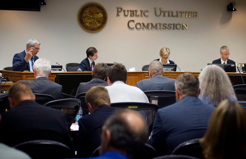 Four people sit at a head table in a meeting room under a sign that says "Public Utilities Commission, with an audience in the foreground.