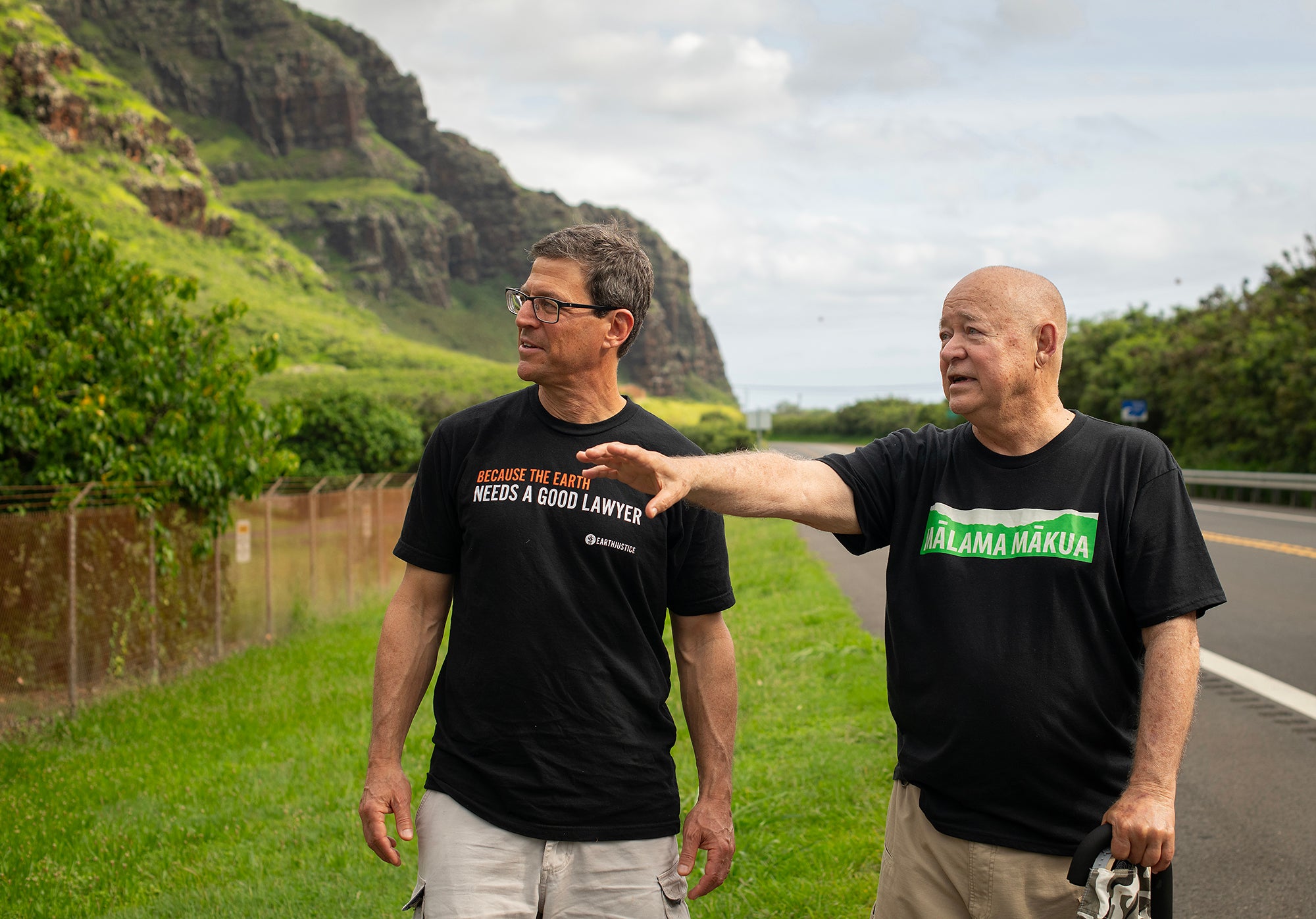 Two men stand between a road and a fence with a large mountain in the background.