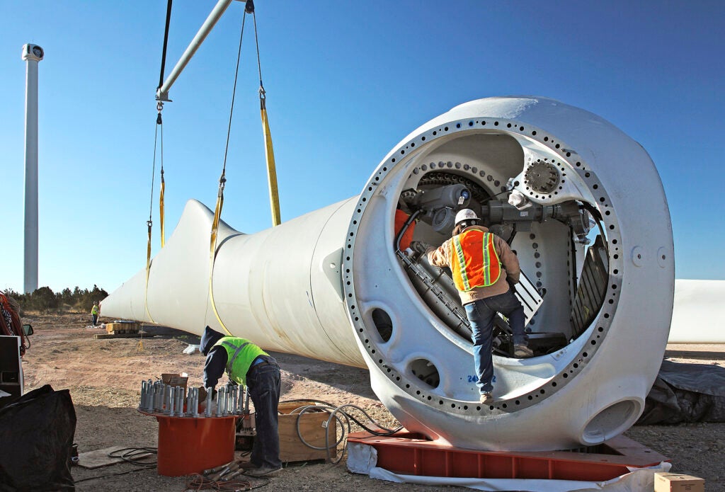 Construction workers prepare a very large wind turbine blade resting on the ground, with the tall turbine tower in the background.