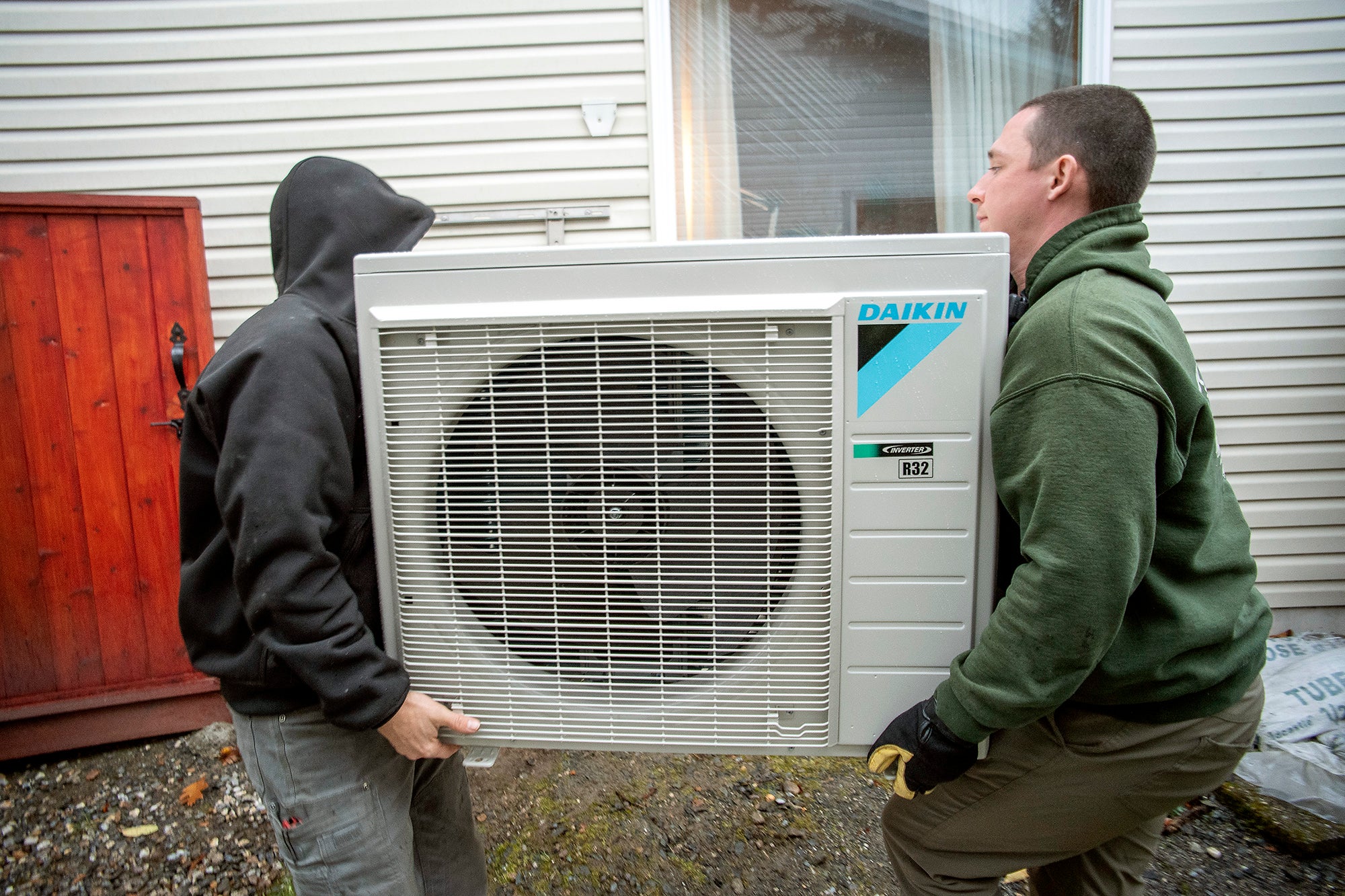 Two men lift a piece of HVAC equipment into place outside of a home.
