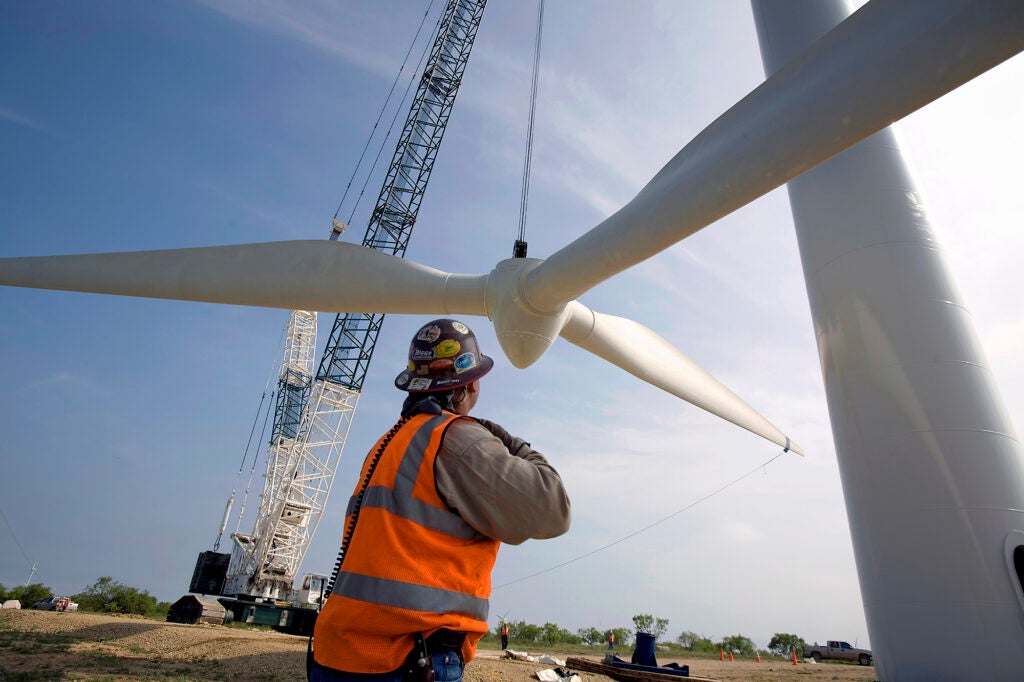 A man in construction gear watches as a very larger turbine blade is lifted into place.
