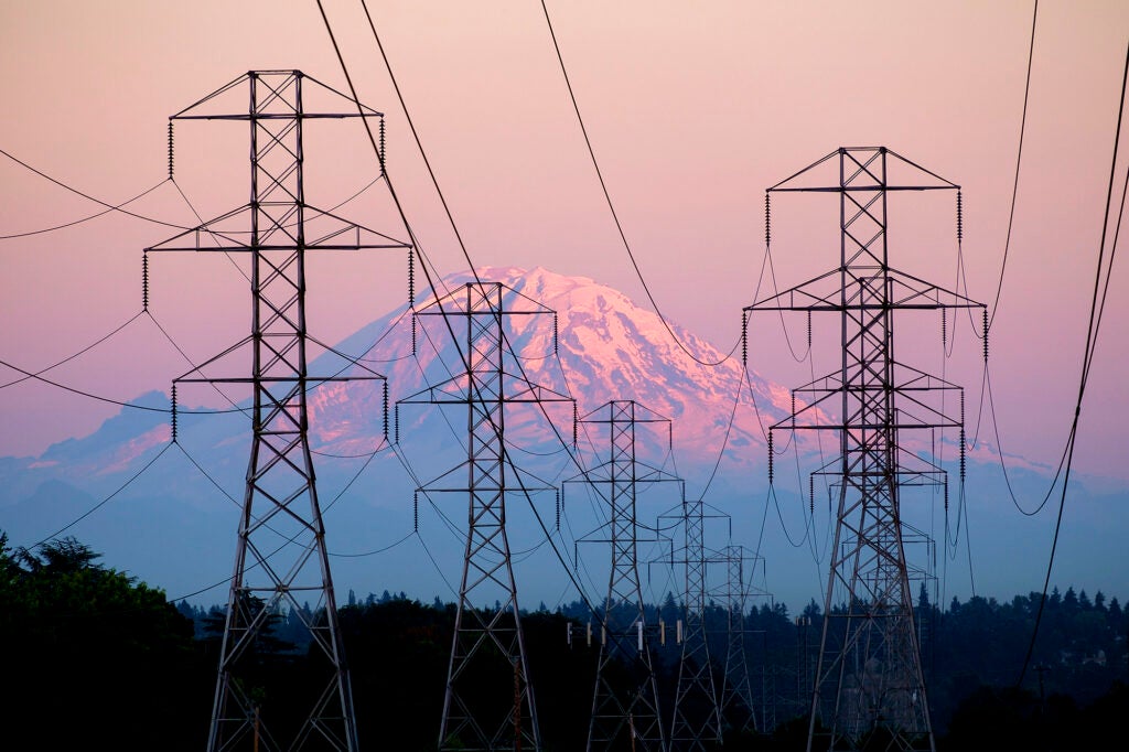Large towers with electrical transmission lines in front of a large mountain at sunset.