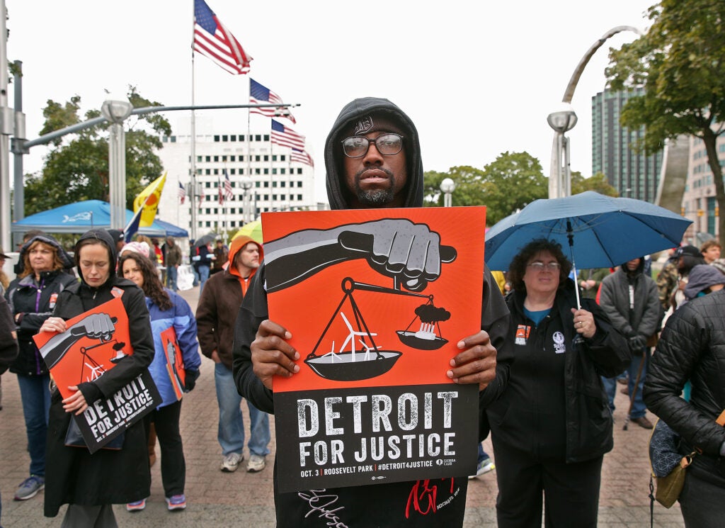 People hold signs saying "Detroit for Justice" with an illustration of scales with wind turbines on one side and a polluting power plant on the other.