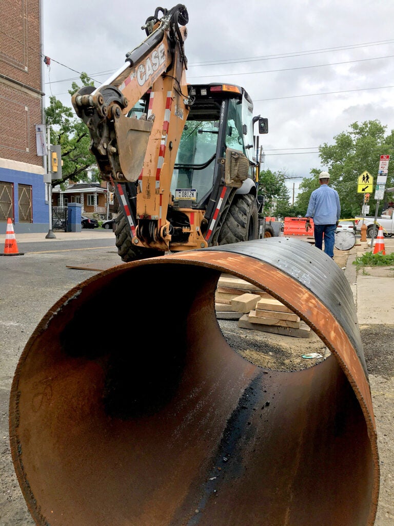 A large metal pipe sits on a city street with an excavator and a man in a hardhat nearby.