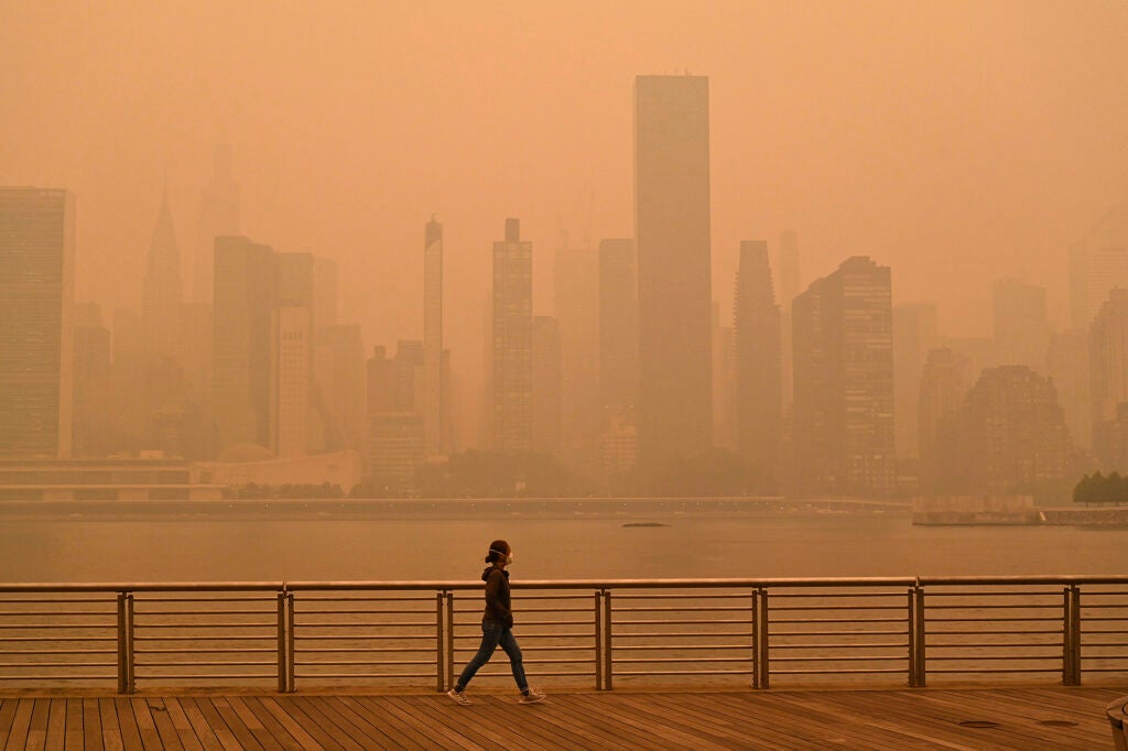 A person wearing a mask walks alone across a river from skyscrapers. Everything is dark and covered in thick orange smoke.
