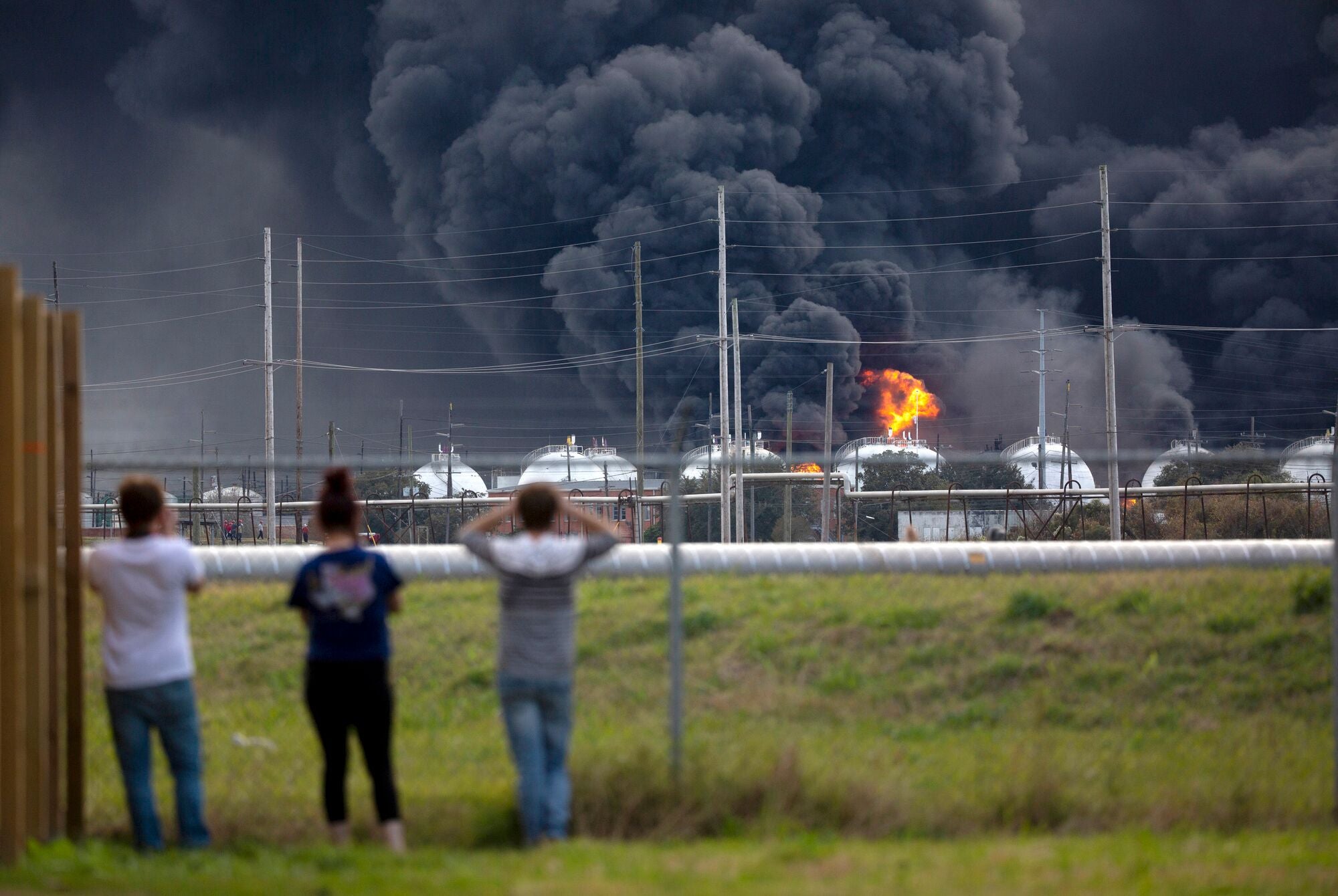 People standing in front of a fence viewing a fire consume a chemical plant