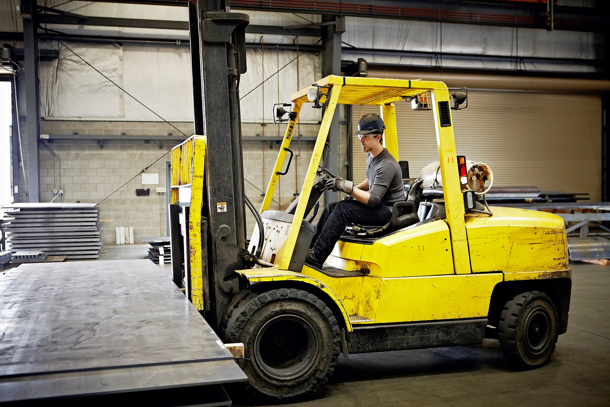 A man wearing a hard hat and safety glasses uses a large yellow forklift to lift large sheets of metal.