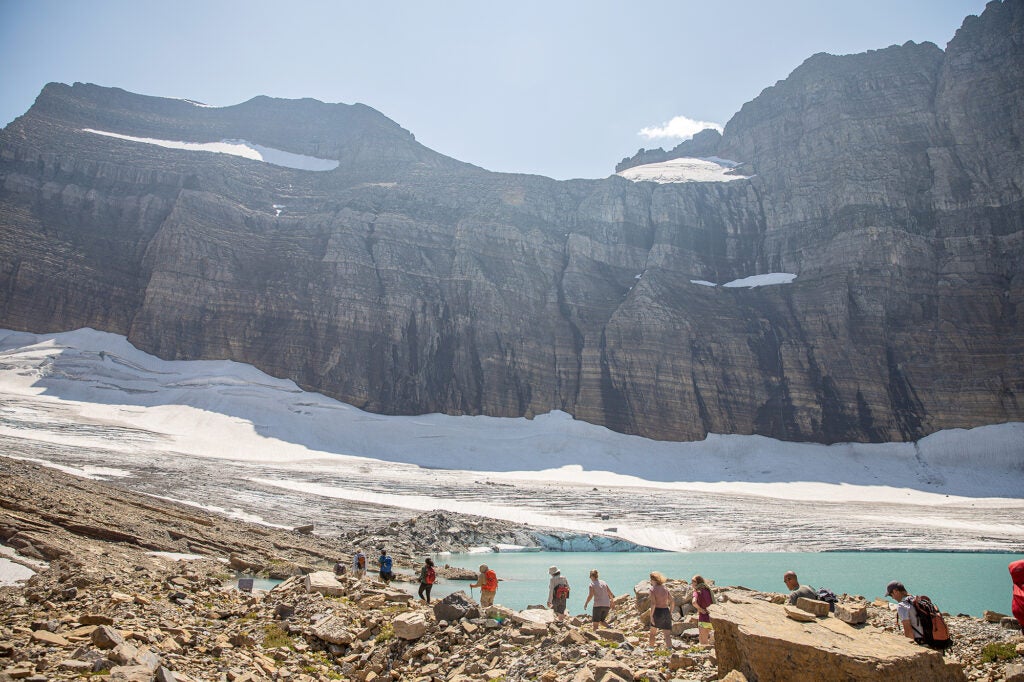 A line of 10 hikers walk towards a distant glacier at the foot of a large cliff, next to a lake.