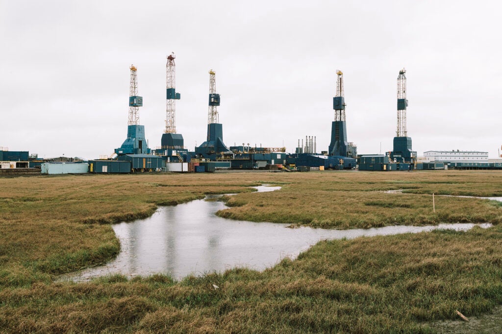 Five drilling towers in a grassy plain with water in the foreground.