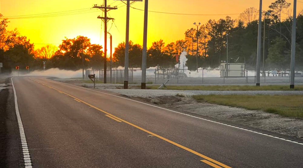 A mist lies low to the ground near a small industrial area, next to a road.