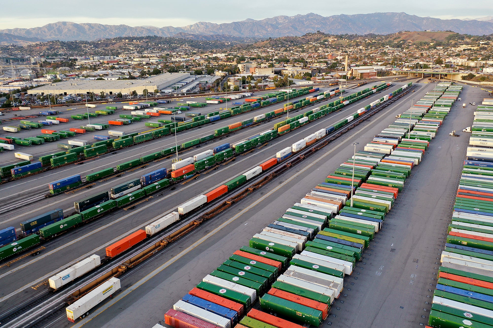 Rows of colorful shipping containers sit in rows and on train cars in a large rail yard next to a commercial and residential part of the city.