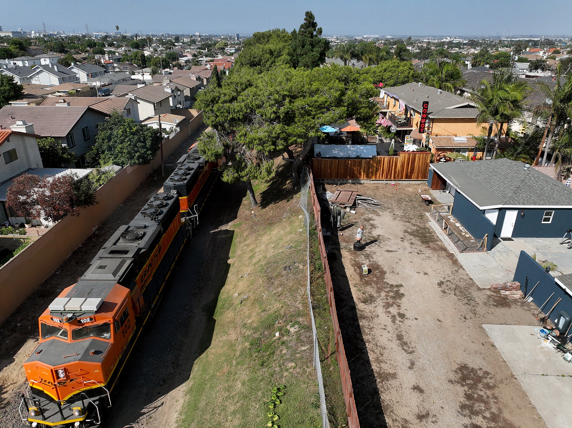 A train with multiple locomotives travels down a small strip of land right next to the backyards of a dense residential area.