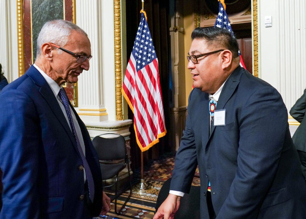 Two men wearing suits stand and talk in an ornate room with American flags behind them.