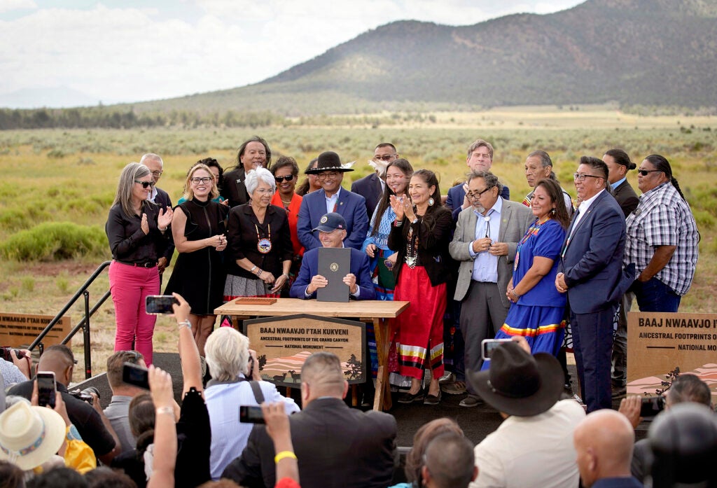 A group of people surrounds a seated Joe Biden, holding up a document. They are in a large plain with a mountain rising behind them.