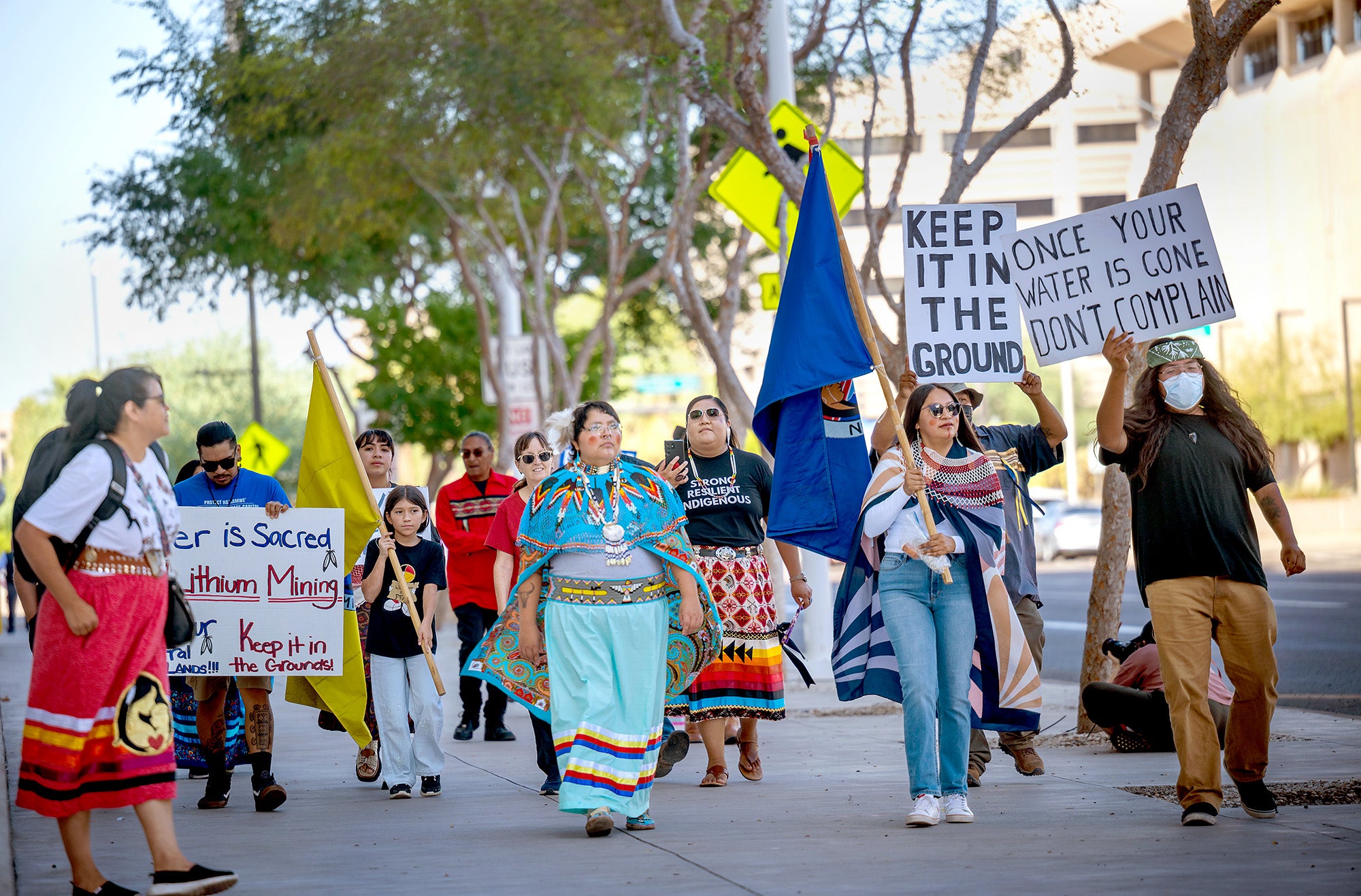 A group of people march on a sidewalk holding flags and signs protesting mining.