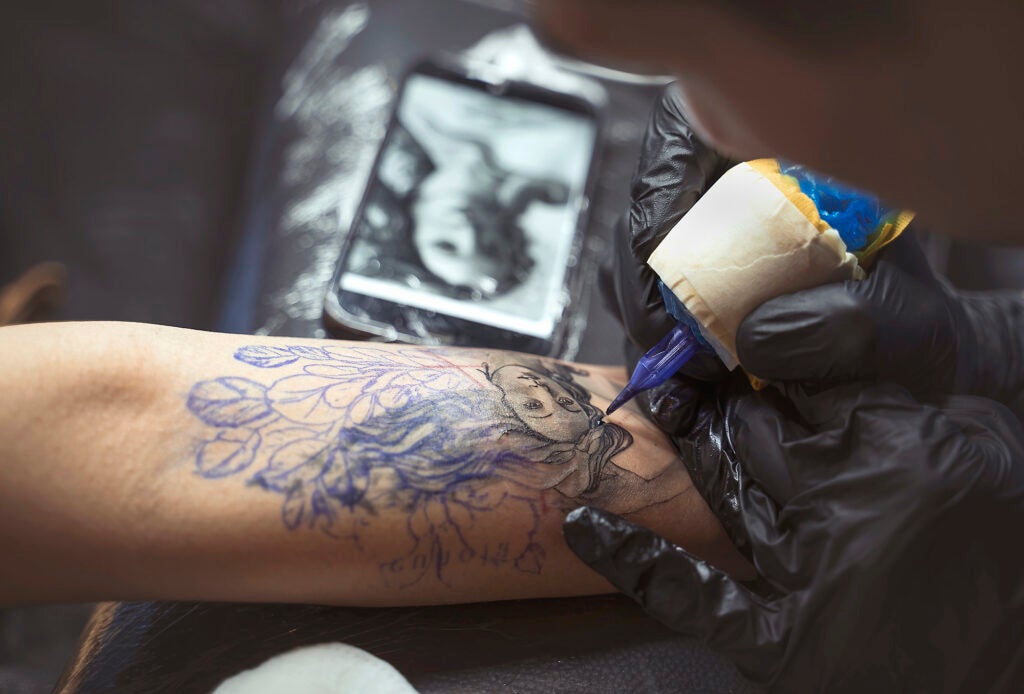 A detail of a tattoo being applied to an arm. The tattoo is a woman's face and hair, which appears to be from the painting "The Birth of Venus"