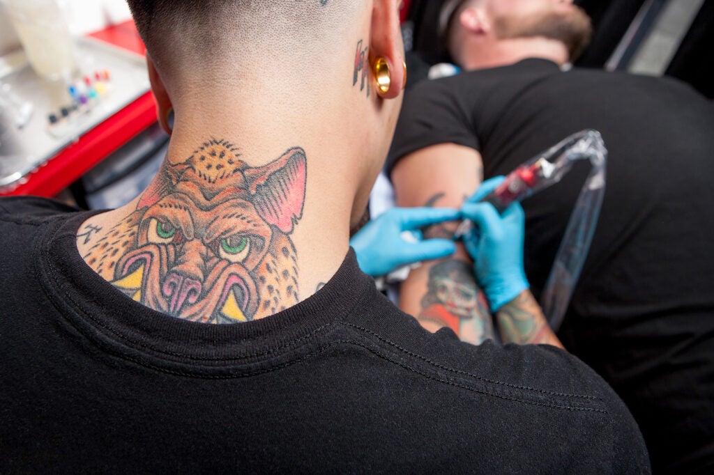 The back of a person's neck with a tattoo in the foreground as the person tattoos another person's arm, which is out of focus.